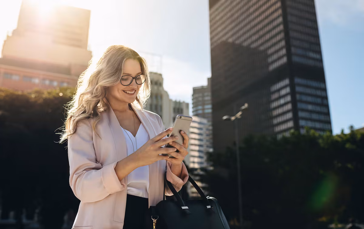 Blonde Frau mit langen Haaren steht draußen vor einem Bürogebäude und schaut lächelnd auf ihr Smartphone.