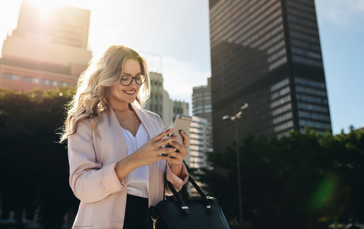 Blonde Frau mit langen Haaren steht draußen vor einem Bürogebäude und schaut lächelnd auf ihr Smartphone.