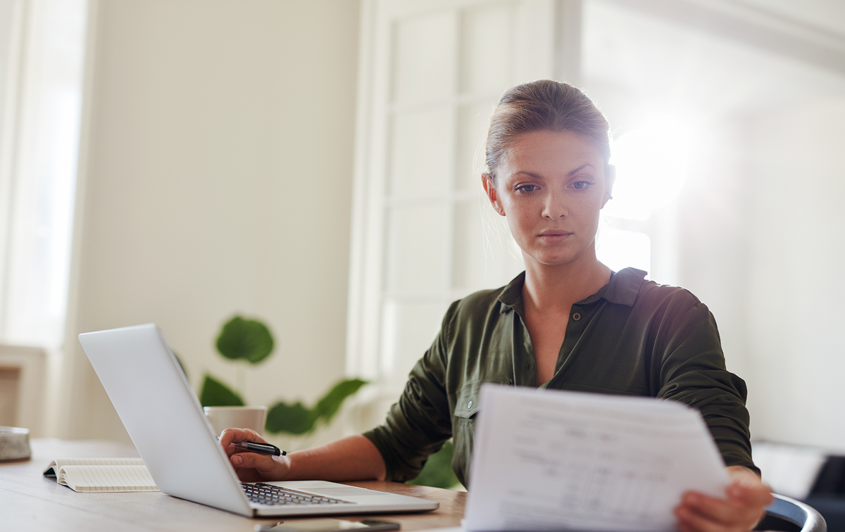 Eine Frau liest ein Dokument am Schreibtisch, während der Laptop aufgeklappt vor ihr steht.