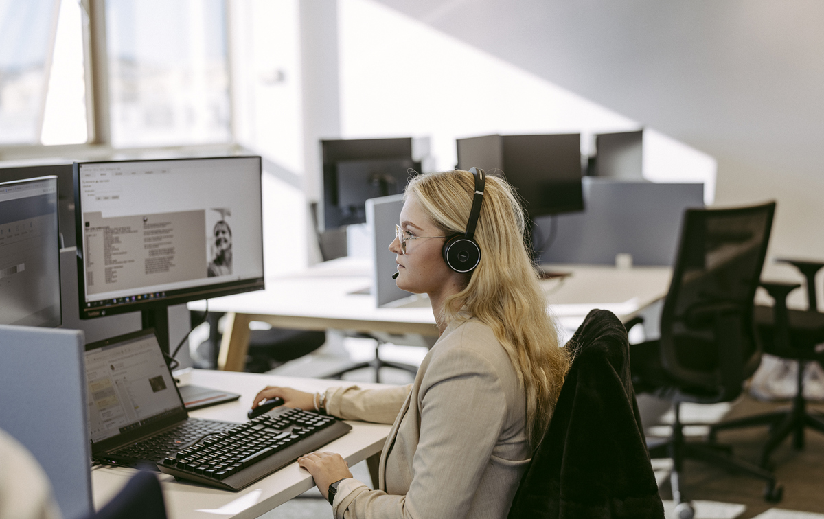 Eine Frau sitzt am Schreibtisch mit einem Headset und arbeitet an einem PC.
